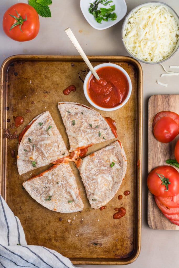 A sliced pizza-dilla with tomato sauce sits on a baking sheet, surrounded by fresh tomatoes, shredded cheese, and a bowl of chopped herbs.