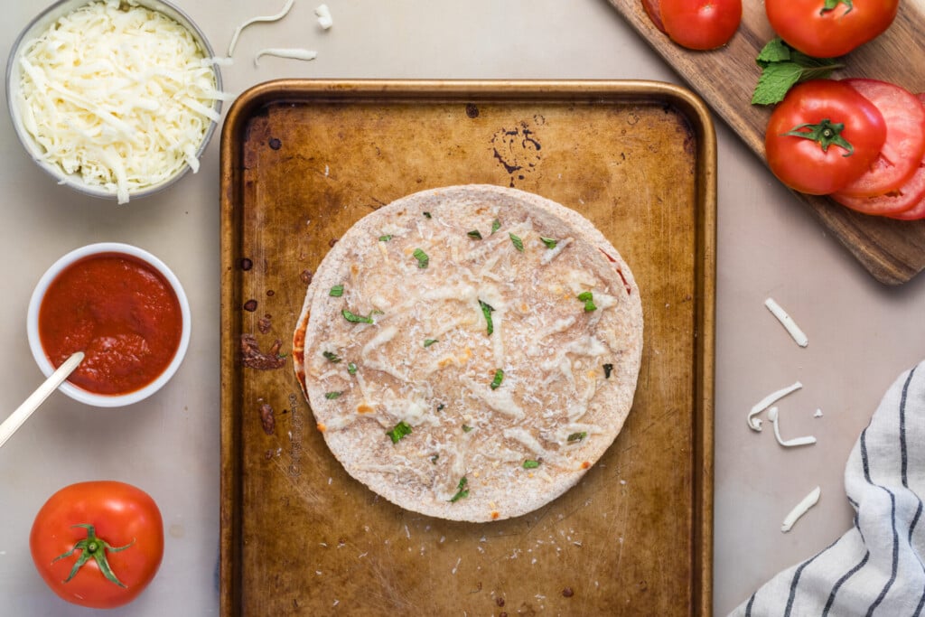 A flatbread pizza-dilla topped with cheese and herbs sits on a metal baking sheet. Surrounding it are a bowl of shredded cheese, a bowl of red pizza sauce, fresh tomatoes on a wooden cutting board, a whole tomato, and a kitchen towel.