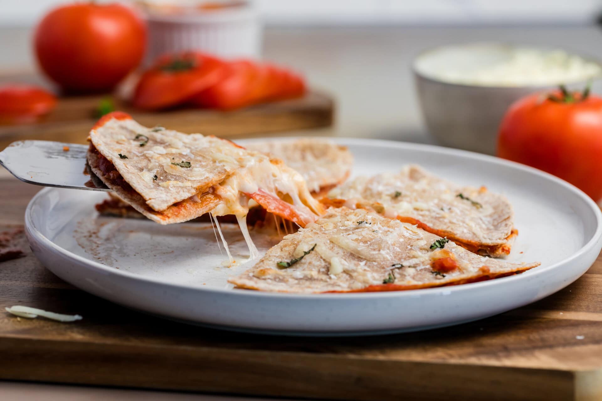 A close-up of a cheesy Pizza-dilla quesadilla being lifted from a white plate, with melted cheese stretching. Fresh tomatoes and a bowl of cheese are in the background.