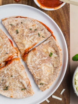 A sliced Pizza-dilla topped with cheese and herbs sits on a white plate, with a bowl of tomato sauce and shredded cheese nearby on a wooden board.
