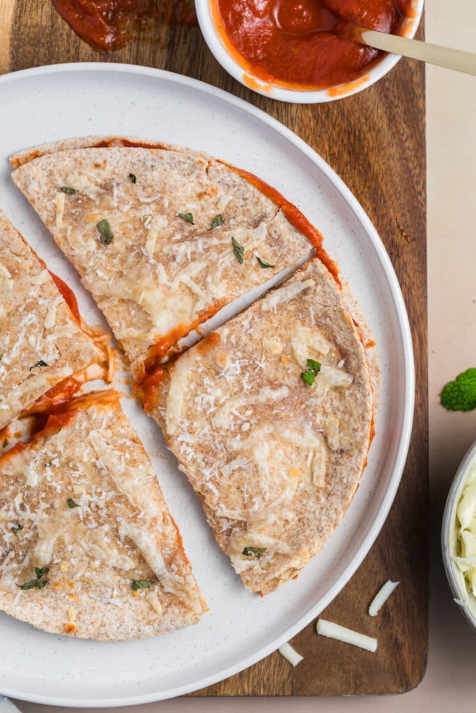 A sliced Pizza-dilla topped with cheese and herbs sits on a white plate, with a bowl of tomato sauce and shredded cheese nearby on a wooden board.