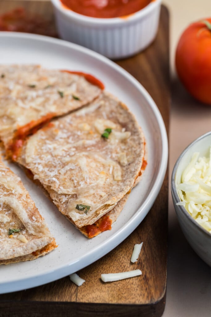 A close-up of a pizza-dilla with cheese and tomato sauce on a plate, resting on a wooden board. A bowl of shredded cheese and a tomato are nearby.