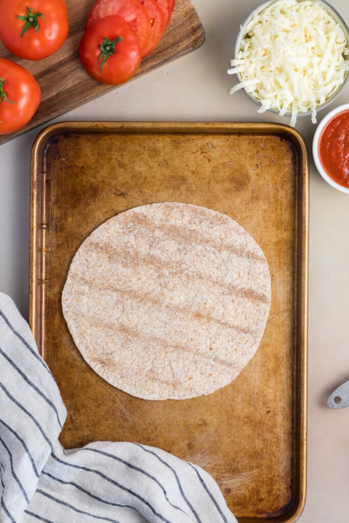 A tortilla on a baking sheet, surrounded by sliced tomatoes, shredded cheese, a bowl of tomato sauce, and a striped cloth—everything you need to create a delicious Pizza-dilla.