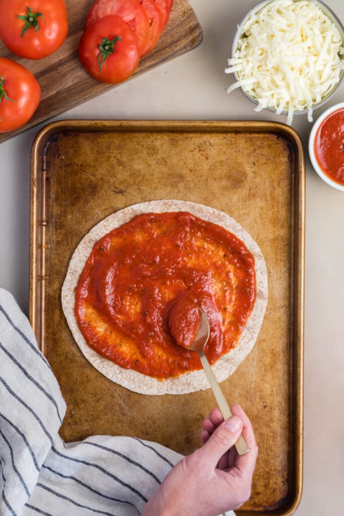 A hand spreads tomato sauce on a round flatbread with a spoon on a baking sheet, preparing the perfect base for a Pizza-dilla. Nearby are sliced tomatoes, shredded cheese, and a bowl of sauce.