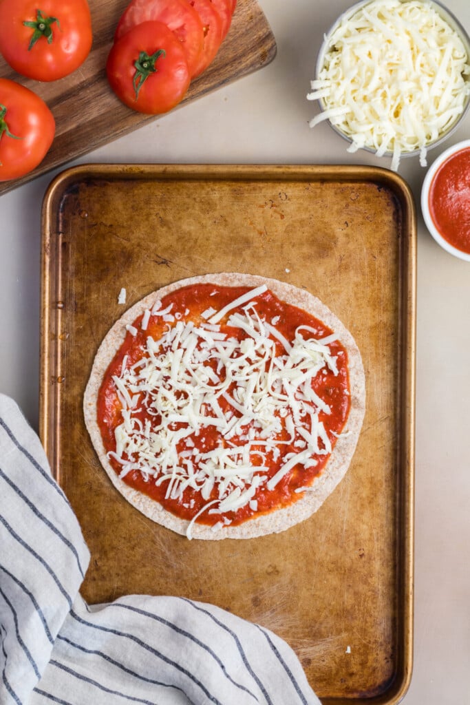 A Pizza-dilla on a baking sheet, featuring a tortilla topped with tomato sauce and shredded cheese. Nearby are a striped kitchen towel, fresh tomatoes, a bowl of shredded cheese, and a bowl of tomato sauce.