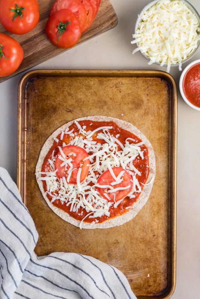 A Pizza-dilla sits on a baking sheet, topped with tomato sauce, shredded cheese, and fresh tomato slices. Nearby are a bowl of shredded cheese, a bowl of sauce, whole and sliced tomatoes, and a striped cloth.