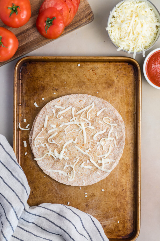 A whole wheat tortilla with shredded cheese on a baking sheet, surrounded by fresh tomatoes, sliced tomatoes, a bowl of shredded cheese, and a bowl of pizza sauce—perfect ingredients for assembling a delicious Pizza-dilla. A striped towel is at the bottom left.