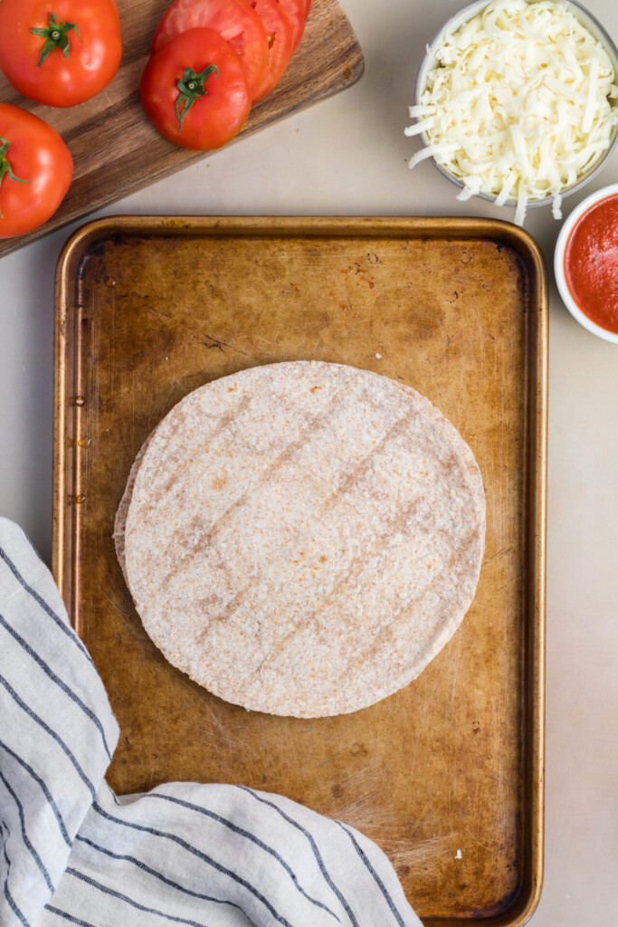 A baking sheet with whole wheat tortillas, perfect for assembling your favorite Pizza-dilla, is surrounded by sliced tomatoes, shredded cheese, tomato sauce, and a striped kitchen towel.