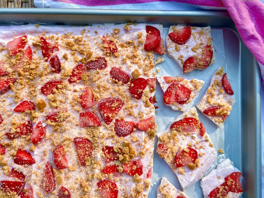 A baking sheet holds a large, rectangular dessert topped with whipped cream, fresh strawberry pieces, and crumbled graham crackers. Several pieces have been cut and separated on the right side. A pink cloth is nearby.
