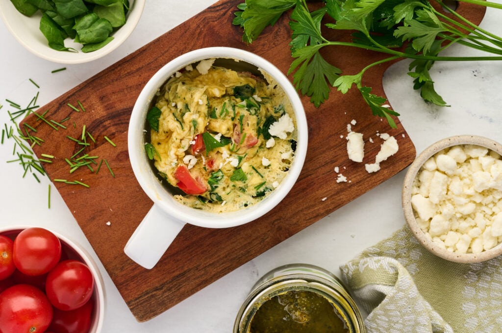 A mug of vegetable omelette sits on a wooden board, surrounded by fresh spinach, parsley, cherry tomatoes, crumbled feta cheese, and a jar of pesto, creating a vibrant and appetizing breakfast scene.