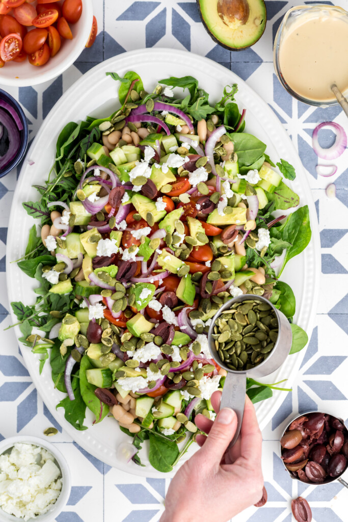 A hand sprinkles pumpkin seeds over a large salad with spinach, avocado, tomatoes, red onion, beans, olives, and crumbled cheese on a white platter. Surrounding bowls hold salad ingredients and dressing.