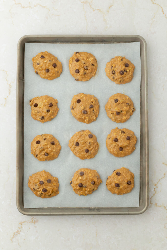 A baking tray lined with parchment paper holds twelve unbaked cookie dough scoops with chocolate chips on a light-colored countertop.