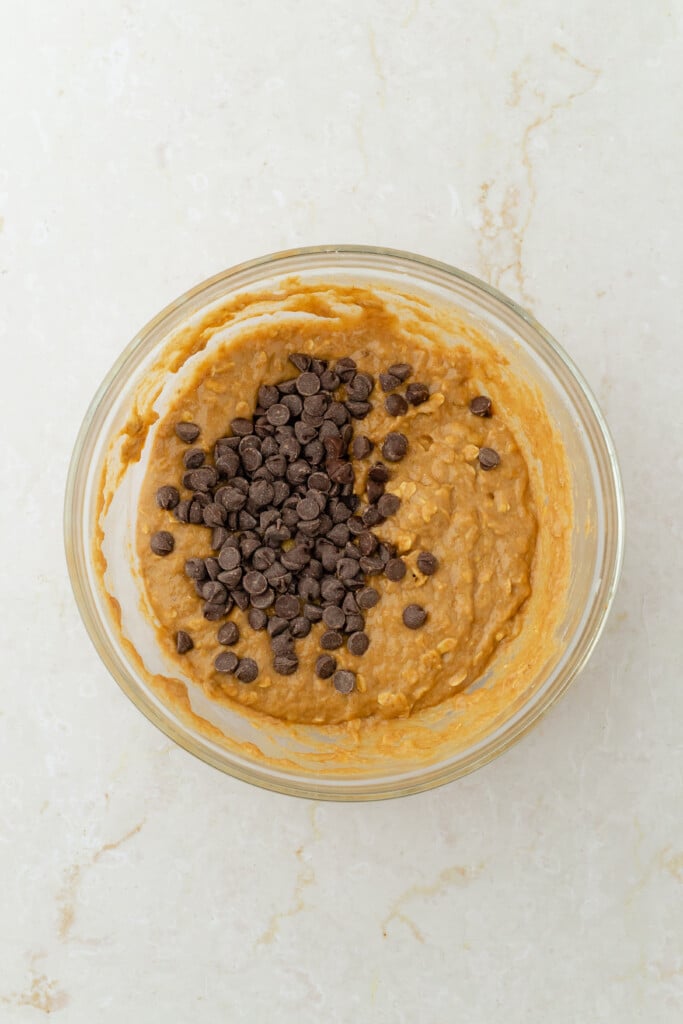 A glass bowl filled with light brown cookie dough topped with a pile of chocolate chips, set on a light-colored, textured countertop.