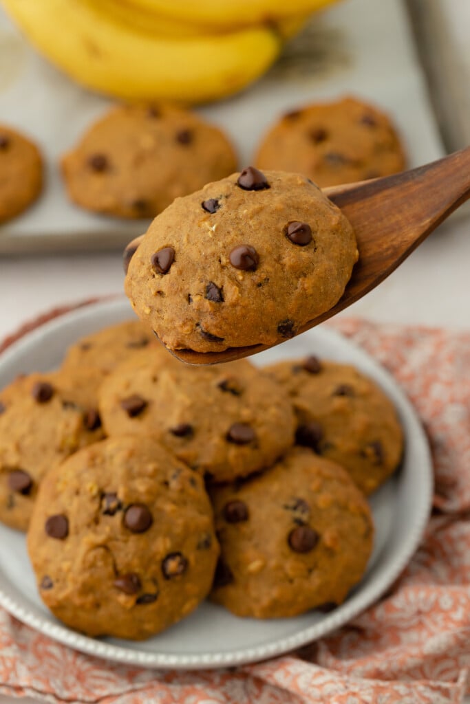 A wooden spoon holds a chocolate chip cookie above a plate stacked with more cookies. Bananas and additional cookies are visible in the background. The cookies appear soft and homemade.