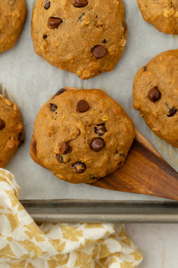 Banana cookies with chocolate chips on a baking tray lined with parchment paper. A wooden spatula lifts one cookie, and a yellow and white patterned cloth is nearby.