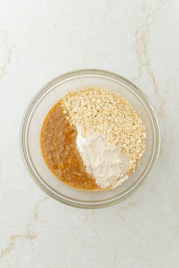 A glass bowl on a light marble surface containing oats, flour, baking powder, and a mixture of wet ingredients, ready to be mixed.