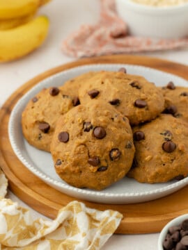 Four chocolate chip cookies sit on a plate with a brown wooden rim. In the background, there are bananas, a bowl of oats, and a patterned napkin, suggesting a cozy, homemade setting.