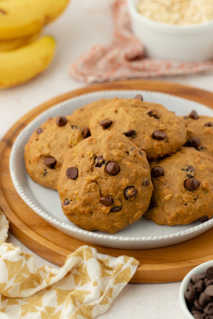 Four chocolate chip cookies sit on a plate with a brown wooden rim. In the background, there are bananas, a bowl of oats, and a patterned napkin, suggesting a cozy, homemade setting.