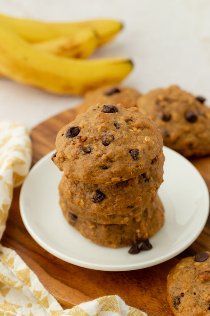 A stack of chocolate chip banana cookies on a white plate, with more cookies in the background. Two ripe bananas and a yellow-patterned cloth are also visible.