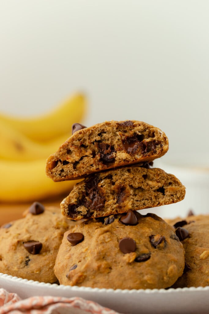 A close-up of two chocolate chip cookies stacked and broken in half, revealing a soft, gooey interior. Whole cookies and chocolate chips are on the plate, with bananas blurred in the background.