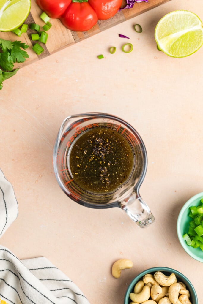 Glass measuring cup filled with sesame ginger dressing for edamame crunch salad, surrounded by fresh vegetables.