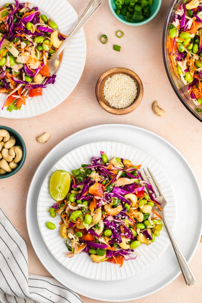 Two plates of edamame crunch salad arranged overhead with sesame seeds, scallions, and cashews nearby.