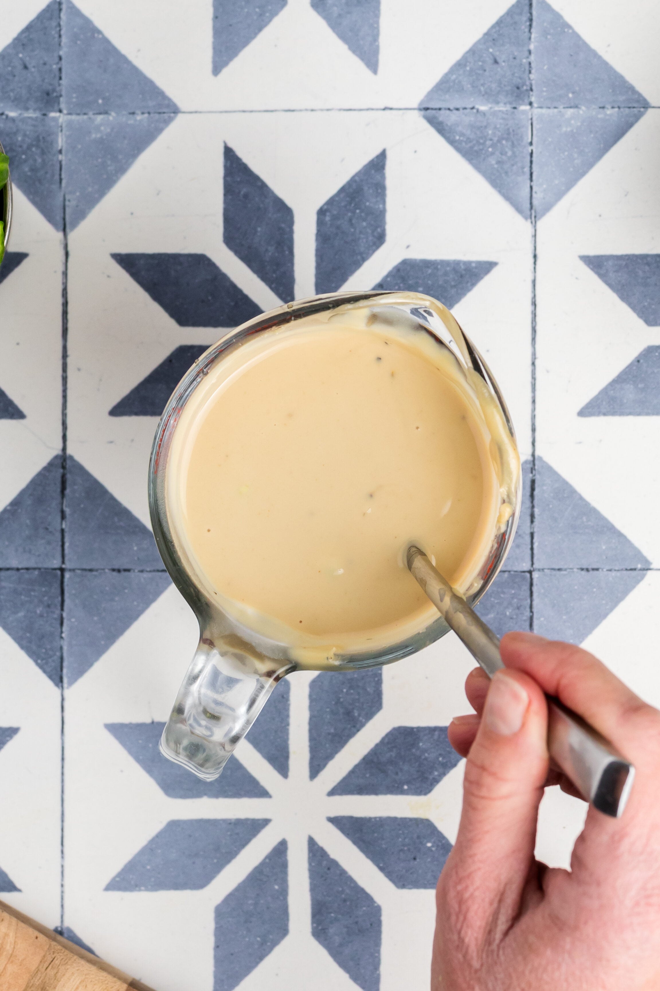 A hand stirs creamy dressing in a glass jug, surrounded by chopped cucumbers, cherry tomatoes, avocado, arugula, feta, red onions, olives, and pepitas on a patterned tile surface.
