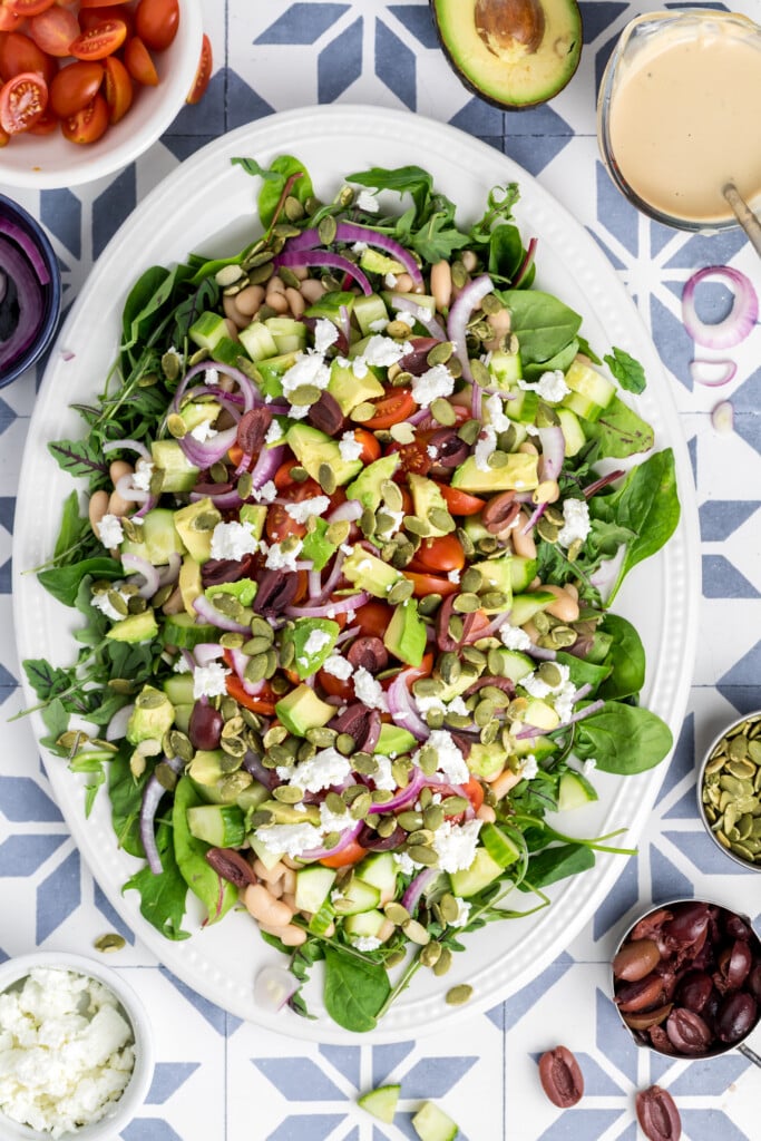 A large oval plate topped with a colorful salad made of spinach, beans, grape tomatoes, red onion, avocado, kalamata olives, pumpkin seeds, and crumbled feta cheese, surrounded by salad ingredients on a blue and white patterned surface.