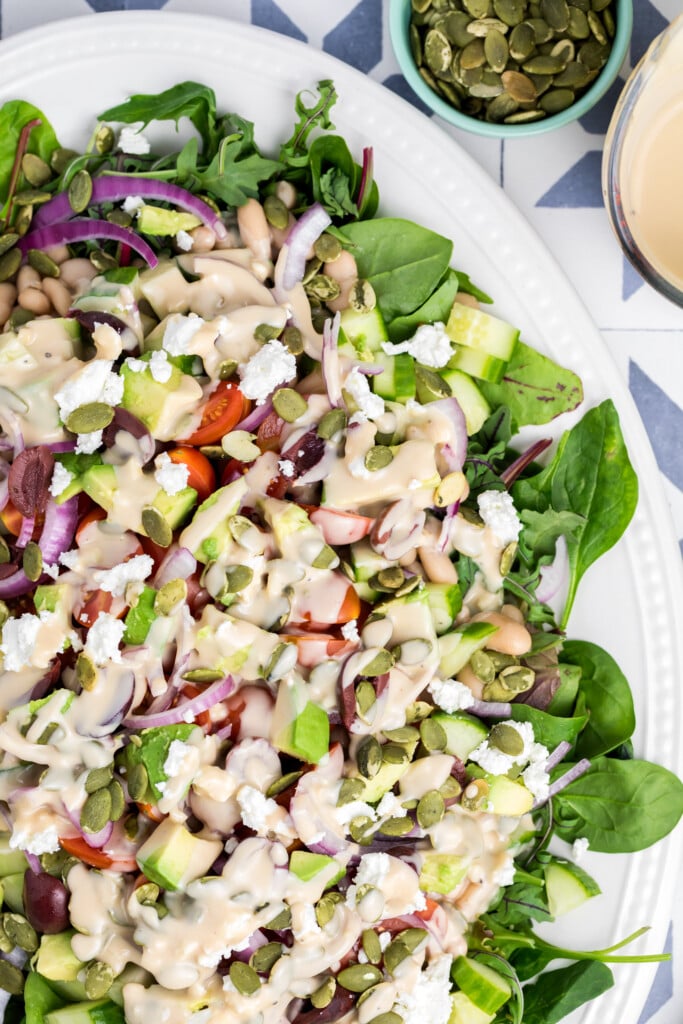 A close-up of a fresh salad with leafy greens, cherry tomatoes, cucumbers, red onions, beans, feta cheese, and pumpkin seeds, drizzled with a creamy dressing on a white plate. A small bowl of pumpkin seeds is nearby.