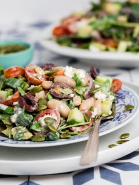 A close-up of a colorful salad with white beans, cherry tomatoes, cucumber, red onion, spinach, feta cheese, olives, and pumpkin seeds on a patterned plate, with a fork resting on the plate.