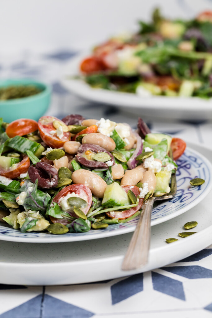 A close-up of a colorful salad with white beans, cherry tomatoes, cucumber, red onion, spinach, feta cheese, olives, and pumpkin seeds on a patterned plate, with a fork resting on the plate.