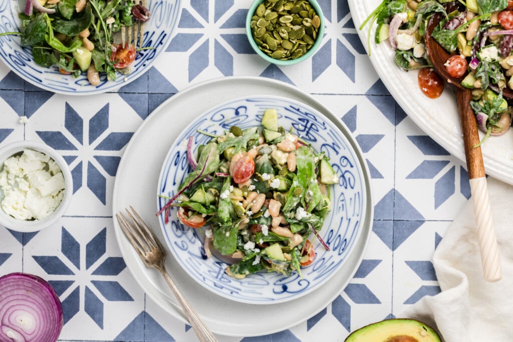 A bowl of fresh salad with green vegetables, cherry tomatoes, cucumbers, nuts and cheese sits on a patterned blue and white tile surface, surrounded by ingredients like pumpkin seeds, avocado, onion and feta cheese.