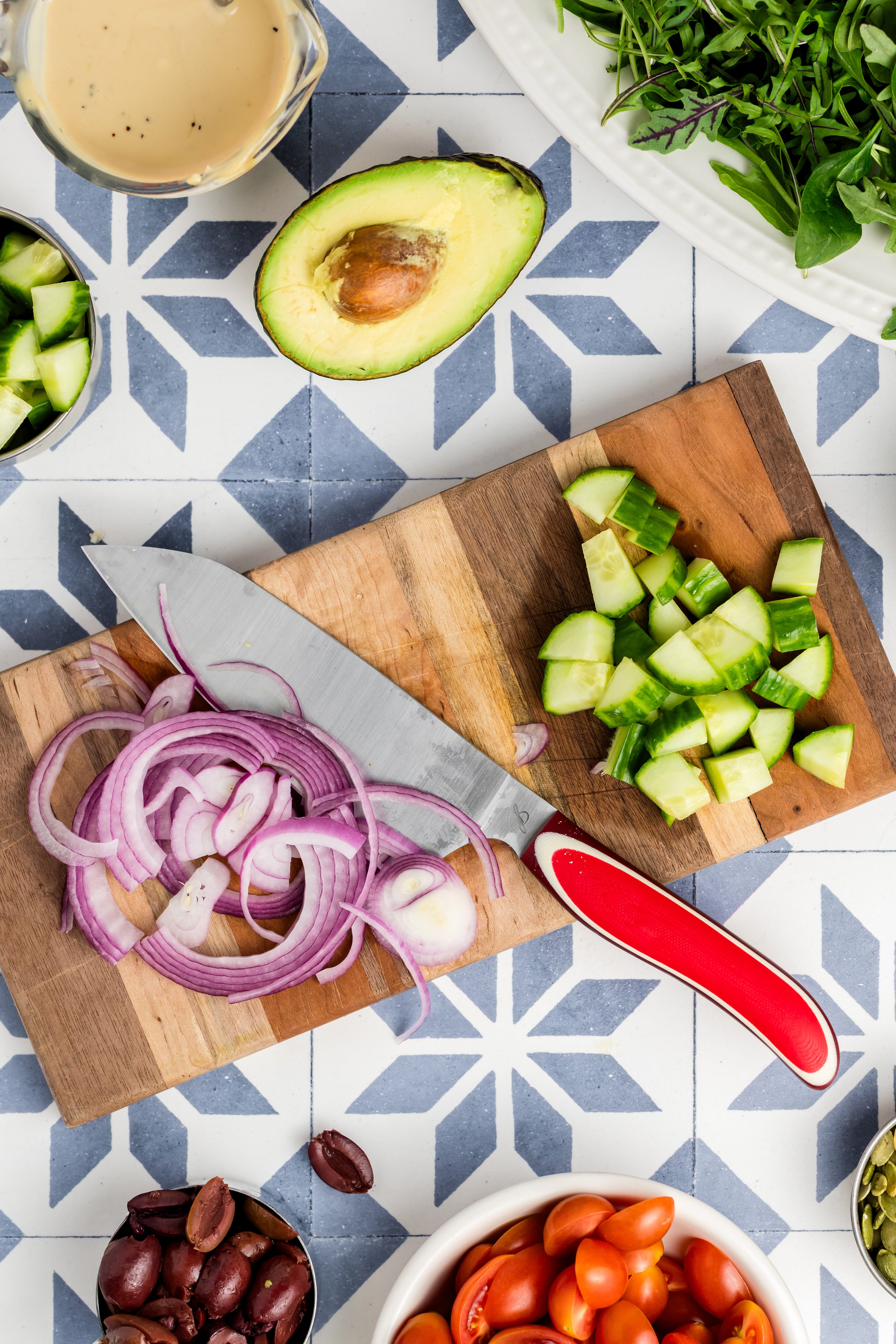 A cutting board with sliced red onion, chopped cucumber, and a knife sits on a patterned surface. Surrounding it are an avocado half, greens, cherry tomatoes, kalamata olives, cucumbers, and a small pitcher of dressing.
