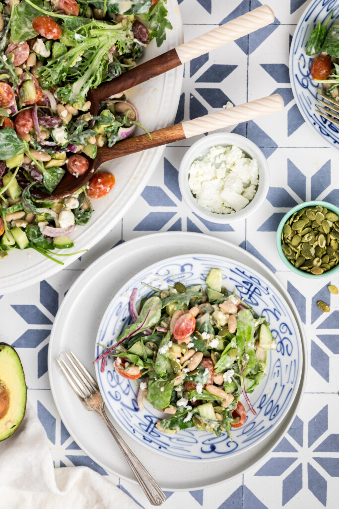 A plate of mixed green salad with cherry tomatoes, avocado, white beans, and cheese, next to a larger salad bowl, a cup of cheese, a cup of pumpkin seeds, and a sliced avocado on a blue and white patterned surface.