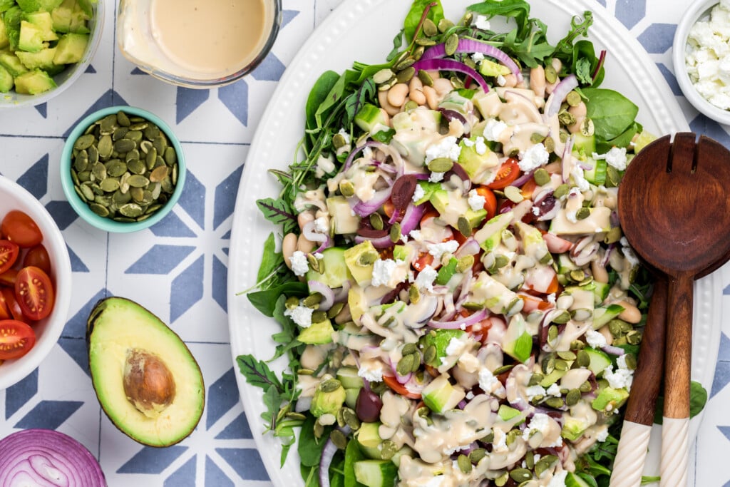 A large white platter of colorful salad with greens, avocado, tomatoes, beans, olives, and cheese, topped with creamy dressing. Surrounding bowls hold salad ingredients on a patterned blue and white table.