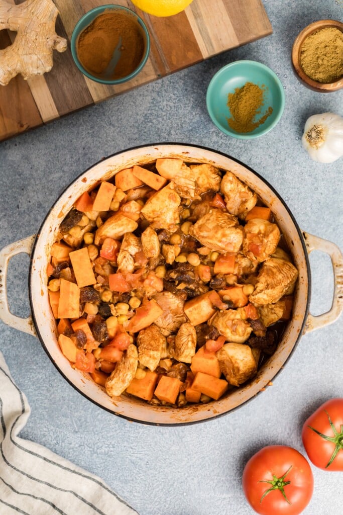 A pot of Moroccan-Spiced Chicken Stew with Sweet Potatoes & Chickpeas sits on a gray surface, surrounded by fresh tomatoes, garlic, ginger, spice bowls, and a striped kitchen towel.