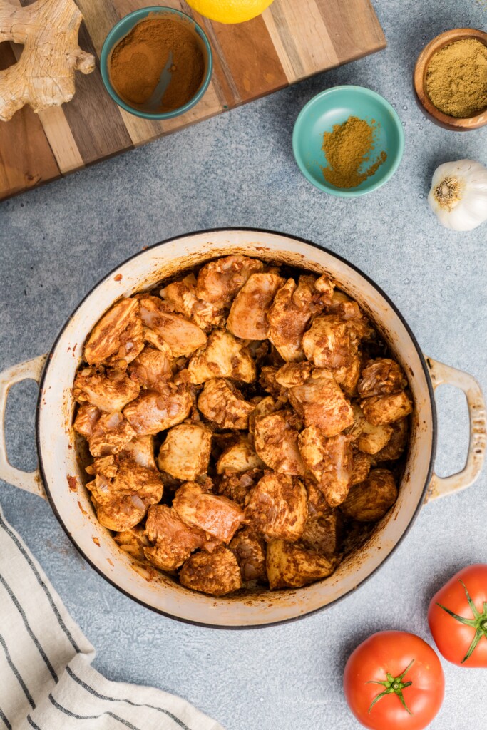 A pot filled with Moroccan-Spiced Chicken Stew with Sweet Potatoes & Chickpeas sits on a countertop, surrounded by fresh tomatoes, a bulb of garlic, bowls of spices, a chunk of ginger, and a striped kitchen towel.