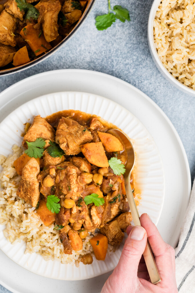 A hand holds a spoon over a plate of brown rice topped with Moroccan-Spiced Chicken Stew with Sweet Potatoes & Chickpeas, garnished with cilantro. Nearby are bowls of curry and rice, plus a sprig of cilantro on a blue surface.