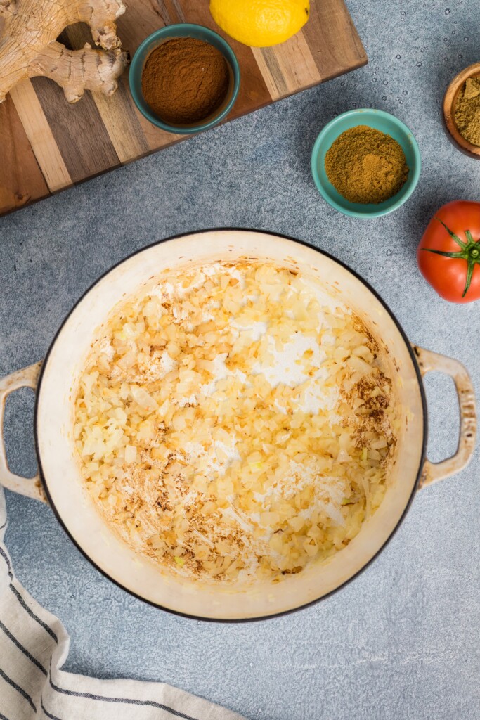 A pot with sautéed chopped onions sits on a gray countertop, ready for Moroccan-Spiced Chicken Stew with Sweet Potatoes & Chickpeas. Nearby are bowls of spices, a tomato, a lemon, ginger, and a striped towel on a wooden cutting board.