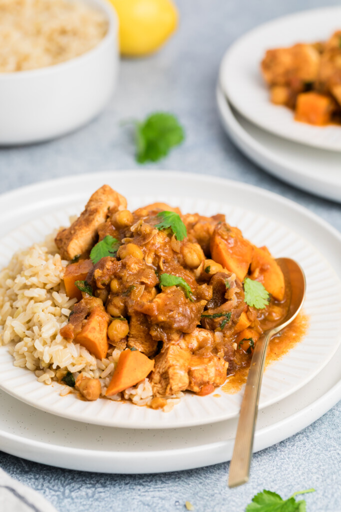 A plate of brown rice topped with Moroccan-Spiced Chicken Stew with Sweet Potatoes & Chickpeas, garnished with fresh cilantro and served with a spoon. Another plate and a bowl of rice are visible in the background.