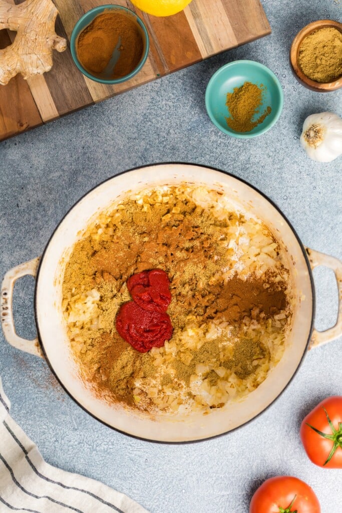 A large white pot holds chopped onions, red tomato paste, and ground spices for Moroccan-Spiced Chicken Stew with Sweet Potatoes & Chickpeas. Nearby are a garlic bulb, fresh tomatoes, and spices, with a wooden cutting board above.