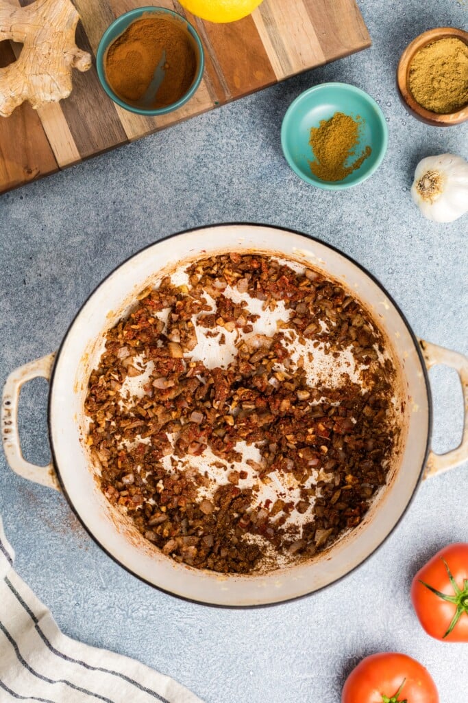 A white enamel pot with sautéed chopped onions, garlic, tomatoes, and spices for Moroccan-Spiced Chicken Stew with Sweet Potatoes & Chickpeas sits on a light blue surface, surrounded by fresh tomatoes, garlic bulb, ginger, and a striped towel.