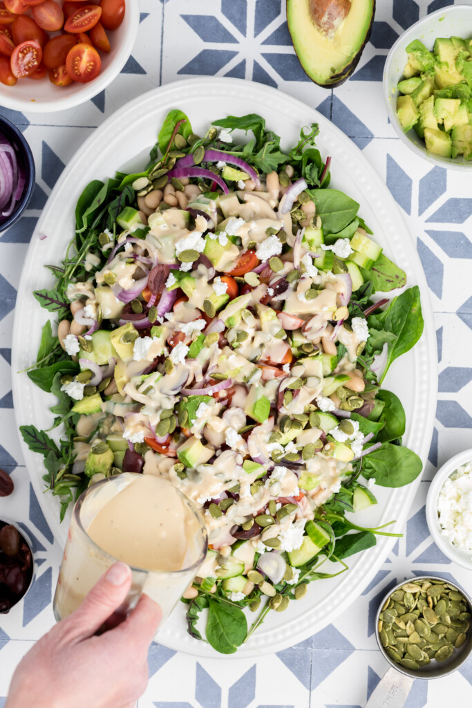 A hand pours creamy dressing over a large platter of salad with spinach, avocado, cucumbers, tomatoes, onions, beans, feta cheese, and seeds on a patterned surface, with salad ingredients arranged around the plate.