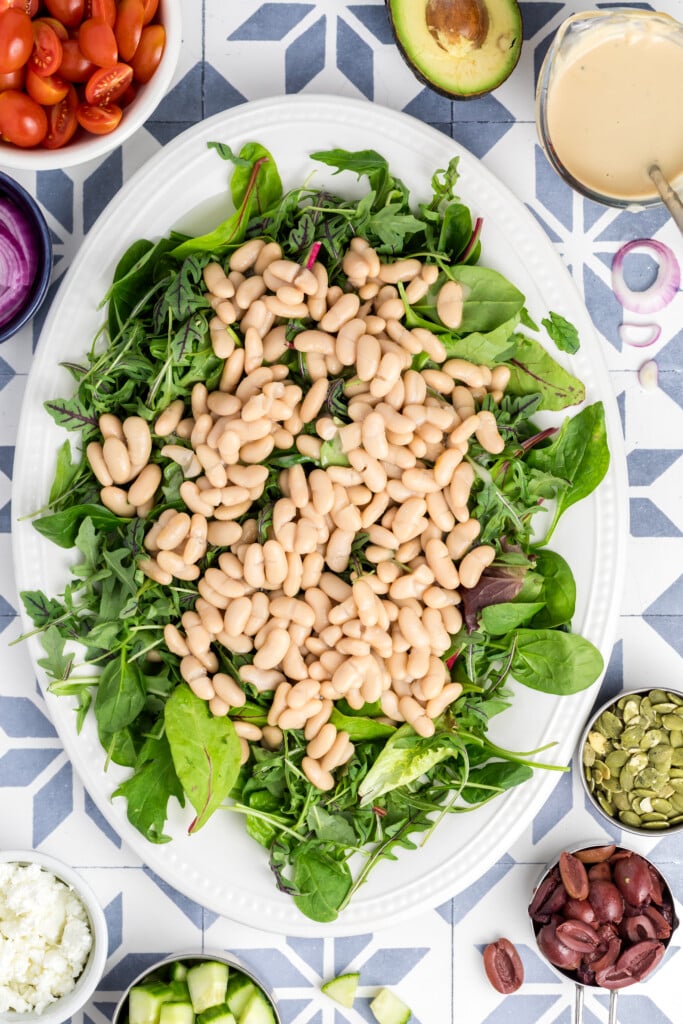 A white platter holds a bed of mixed greens topped with white beans. Surrounding the platter are bowls of cherry tomatoes, avocado, dressing, red onions, pepitas, olives, cucumber, and feta cheese on a patterned surface.