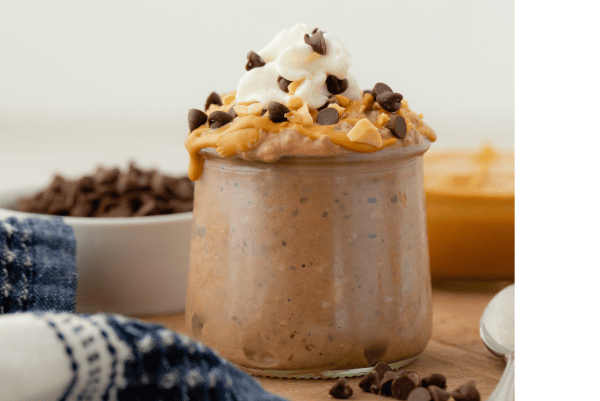 A glass jar filled with chocolate peanut butter overnight oats, topped with whipped cream, chocolate chips, and chopped peanuts—one of the best overnight oat recipes for a decadent breakfast. A bowl of chocolate chips and a spoon are nearby.