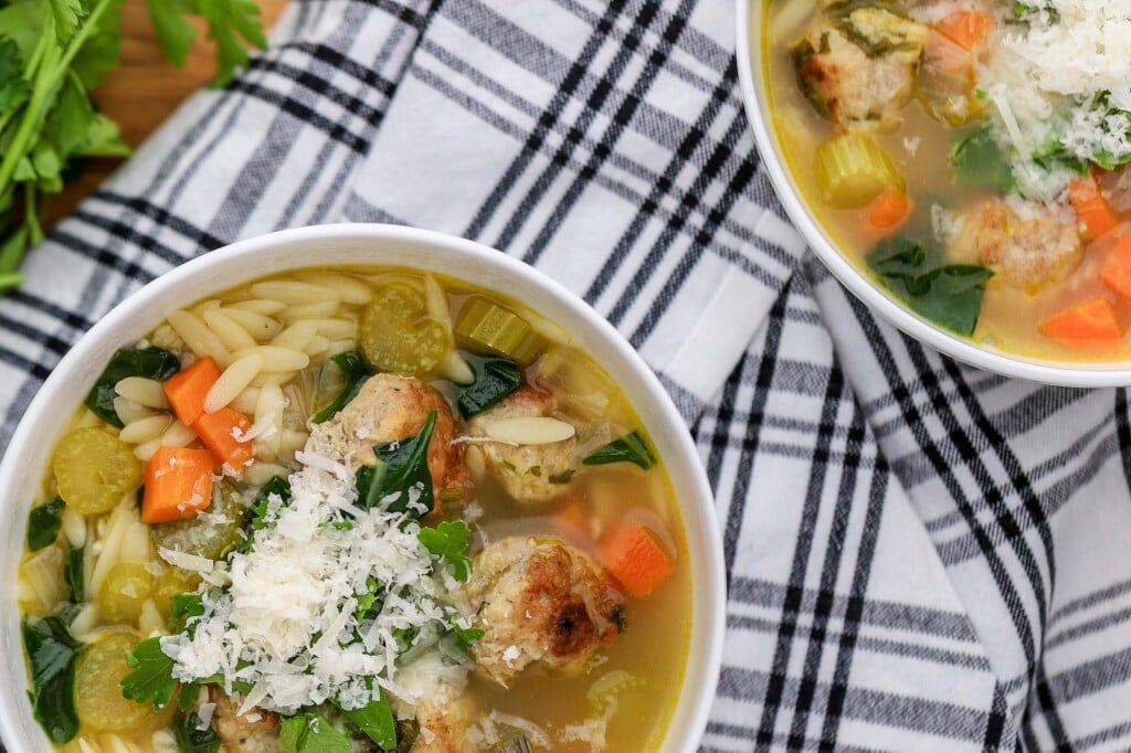 A bowl of One-Pot Italian Wedding Soup with meatballs, orzo pasta, carrots, celery, spinach, and grated cheese sits on a black-and-white plaid cloth. Fresh parsley is on a wooden board beside the bowl.