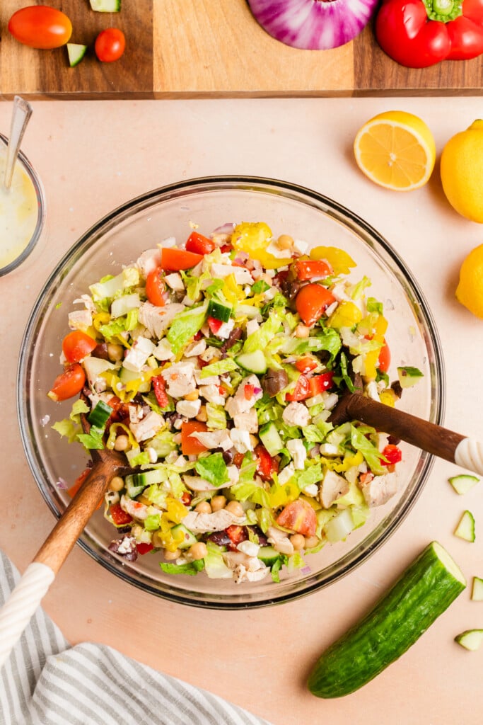 A glass bowl of chopped salad with tomatoes, cucumbers, olives, lettuce, and feta cheese is being mixed with wooden utensils. Surrounding the bowl are fresh vegetables, lemon halves, and a small bowl of dressing.