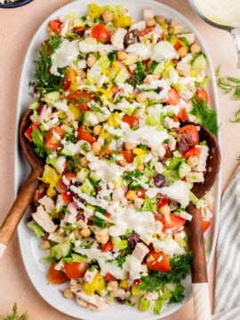 A large white platter filled with a colorful chopped salad including lettuce, tomatoes, cucumber, chickpeas, olives, peppers, chicken, and herbs, topped with a creamy white dressing, with wooden salad servers on the sides.