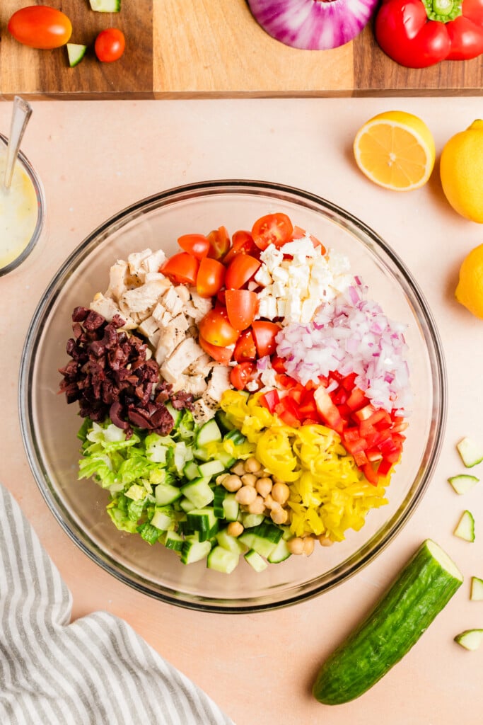 A glass bowl filled with salad ingredients: chopped lettuce, cucumbers, chickpeas, kalamata olives, grilled chicken, grape tomatoes, feta cheese, red onion, red bell pepper, and pepperoncini, seen from above.