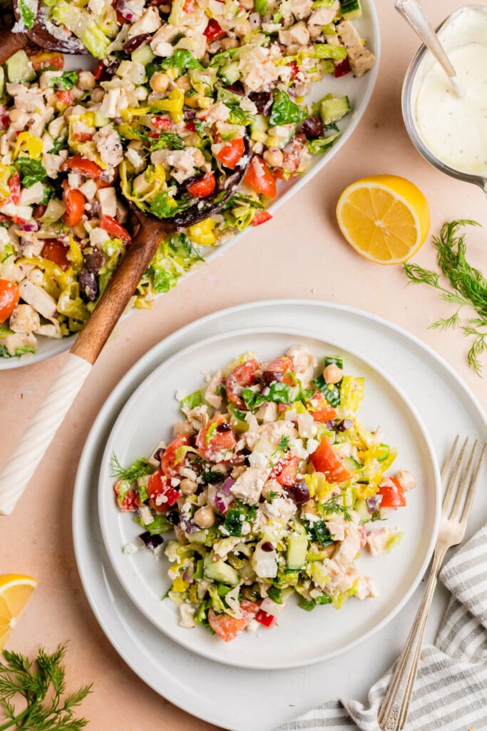 A plate of chopped salad with lettuce, tomatoes, cucumbers, red onions, chickpeas, feta cheese, and herbs. A large bowl of salad, sliced lemon, fresh dill, and a fork are nearby on a light pink surface.
